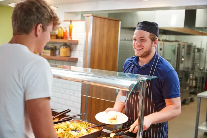 A student being served a meal by a staff member at a campus dining hall serving line.