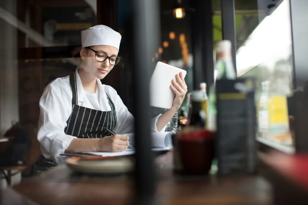 A chef in a white uniform writing notes beside a tablet.