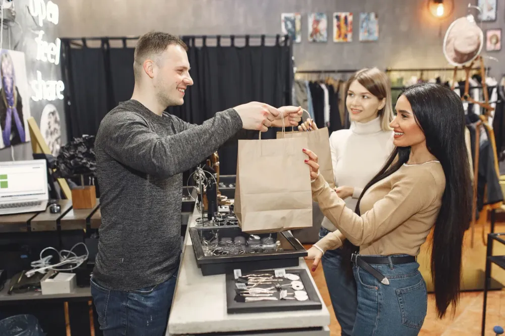 A store associate handing a paper shopping bag to two customers at a counter.