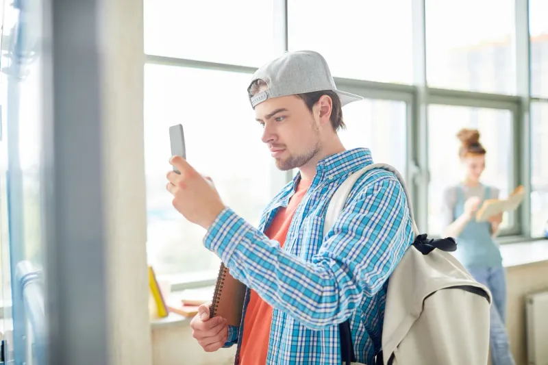 A student with a backpack holding up his phone in a bright school corridor.