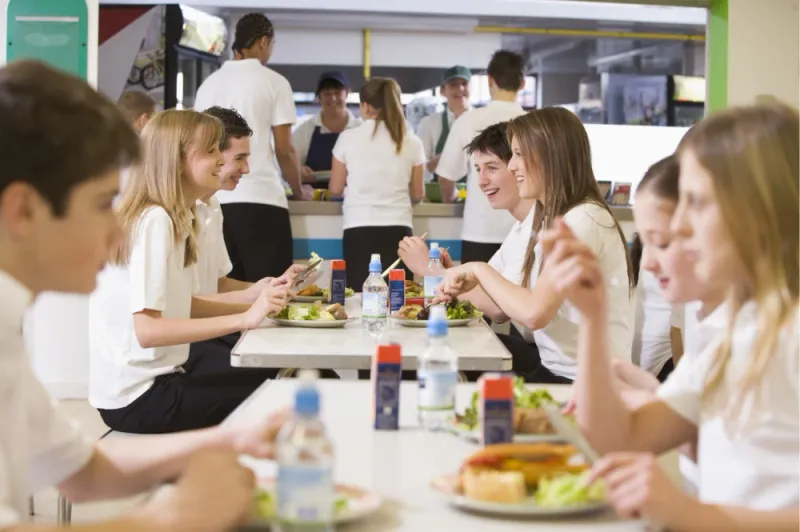 Students eating lunch together in a school cafeteria while staff serve food behind them.